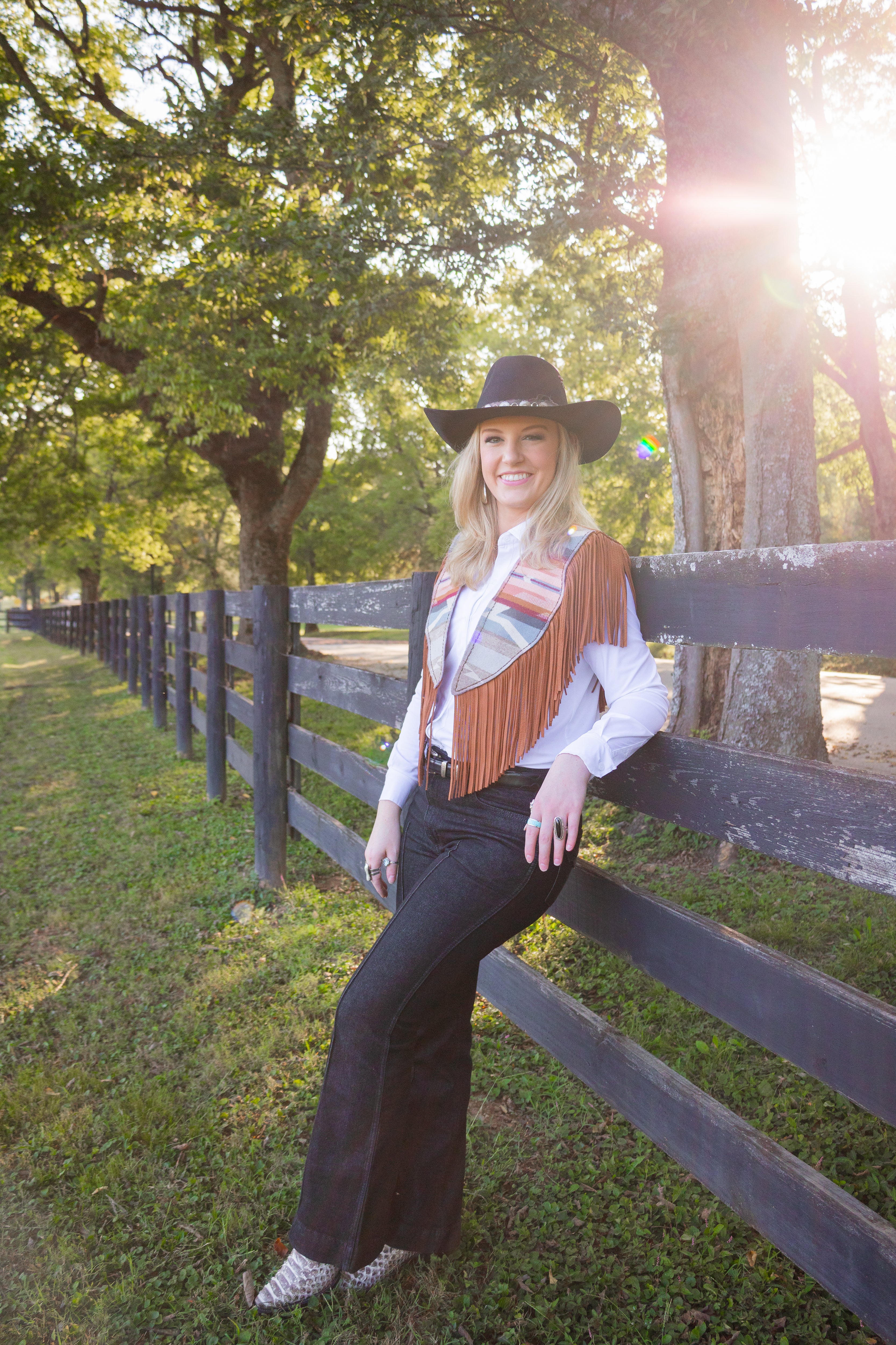 Woman in cowboy hat and vest sitting on a wooden fence with trees and sunlight in the background