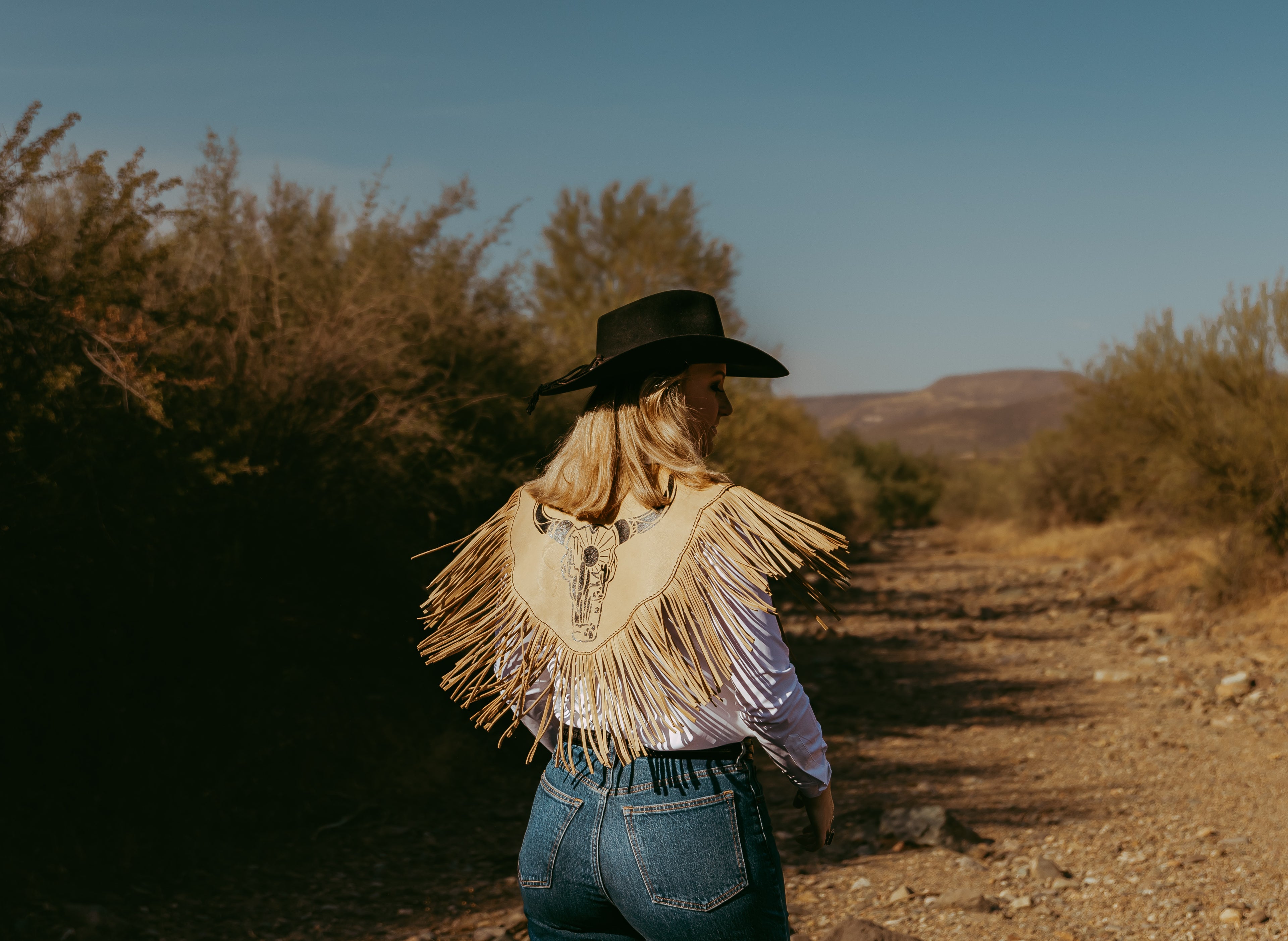Person in a cowboy hat and fringed jacket walking on a dirt path in a desert landscape.